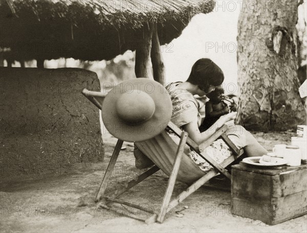 Mollie with Sudanese baby. A European woman captioned as 'Mollie' plays with a Sudanese baby sitting on her knee in a deckchair beside a mud hut. Her hair is stylishly cut into a short bob and her wide brimmed hat hangs casually from the back of her chair. Sudan, circa 1930. Sudan, Eastern Africa, Africa.