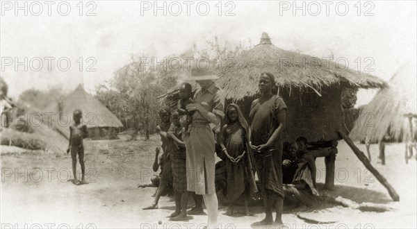 Mollie with a Golo family. A European woman captioned as 'Mollie' smiles down at the Sudanese baby she is holding. The baby's family stand nearby outside a mud hut in their home village. Golo, Sudan, circa 1930. Golo, West Darfur, Sudan, Eastern Africa, Africa.