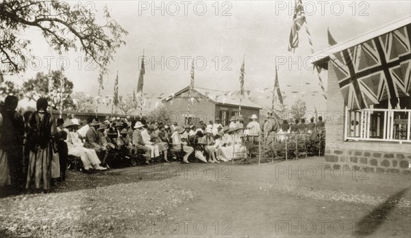 Opening of the Stack Memorial School. Two African priests stand in the shade of a tree beside a seated European crowd at a ceremony held for the opening of the Stack Memorial School. The colonial-style buildings are decorated with union jack flags and bunting. Wau, Sudan, 1927., West Bahr el Ghazal, Sudan, Eastern Africa, Africa.