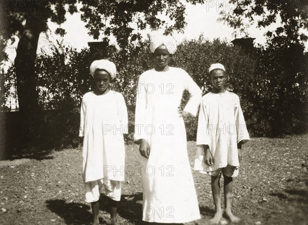 Mess servants. Three African mess servants, an adult male and two boys, stand side by side in a garden wearing turbans and white robes. Sudan, circa 1925. Sudan, Eastern Africa, Africa.