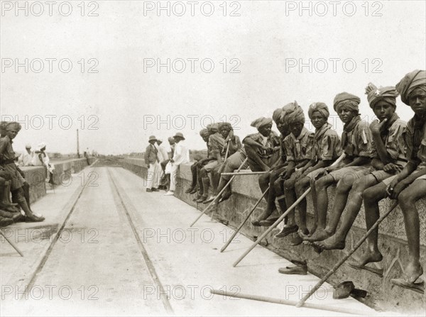 Scout group on the Sennar Dam. An African scout group sit on top of the nearly completed Sennar Dam. Each carries a stick and wears a uniform consisting of short trousers, a shirt and neckscarf. Sennar, Sudan, circa 1925. Sennar, Sennar, Sudan, Eastern Africa, Africa.