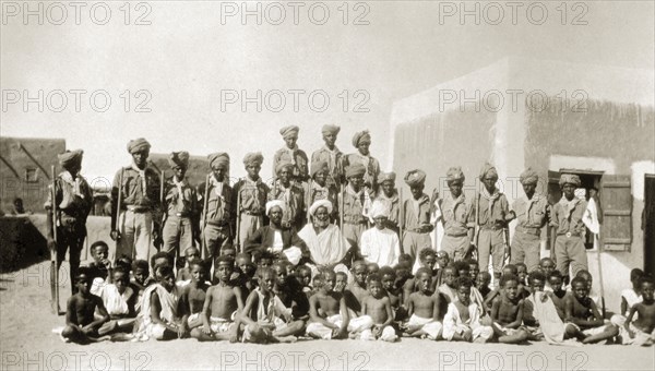 African scout group. Portrait of an African scout group in Sinkat. Uniformed scouts carrying sticks stand in line behind a large group of boys sat clustered together on the floor. Three African officials wearing robes and turbans sit in the centre of the group. Sinkat, Sudan, 1926. Sinkat, Red Sea, Sudan, Eastern Africa, Africa.