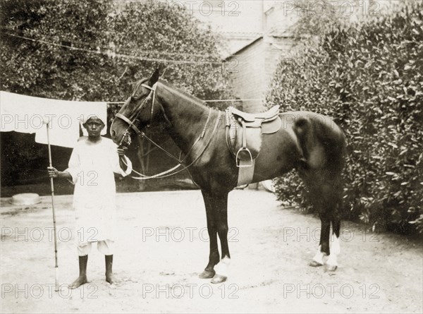 Tinto the horse. A young African servant stands polo mallet in hand, displaying a horse captioned as 'Tinto' who is saddled up ready for a game of polo. Sudan, circa 1925. Sudan, Eastern Africa, Africa.