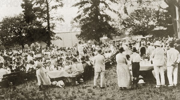 Naval lunch at Pietermaritzberg. A large crowd of men and boys dressed in white naval uniforms sit down around long tressle tables outdoors waiting to eat their lunch. Pietermaritzberg, Natal (KwaZulu Natal), South Africa, 1-5 January 1924. Pietermaritzburg, KwaZulu Natal, South Africa, Southern Africa, Africa.