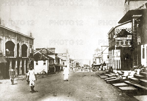 Sierra Leone street scene. A handful of people mill about on a wide road in the centre of a large town. The road is flanked by buildings of different styles and leads down towards the sea. Sierra Leone, 8-13 December 1923. Sierra Leone, Western Africa, Africa.