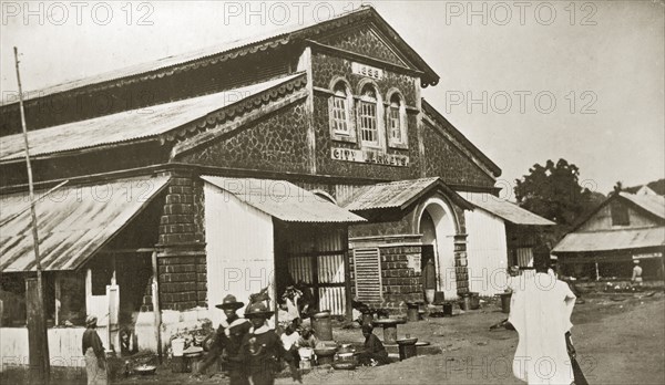 City market, Sierra Leone. A handful of people mill about outside a large stone market building in the centre of a town. Signs carved in stone above the front entrance read: 1898, CITY MARKETS. Sierra Leone, 8-13 December 1923. Sierra Leone, Western Africa, Africa.