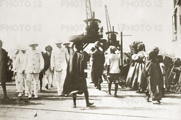 Landing stage. An African man carrying a basket of bananas on his head, weaves through a crowd at a landing stage. European men to his left wear white naval uniforms: female African workers to his right stand beside crates of bananas. Sierra Leone, 8-13 December 1923. Sierra Leone, Western Africa, Africa.