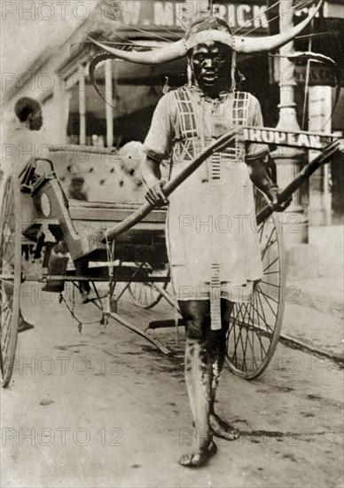 Rickshaw puller in Pietermaritzberg. A rickshaw puller stands between the shafts of his empty carriage on a main street, barefoot and wearing a flamboyantly horned and feathered headdress. Pietermaritzberg, Natal (KwaZulu Natal), South Africa, 1-5 January 1924. Pietermaritzburg, KwaZulu Natal, South Africa, Southern Africa, Africa.