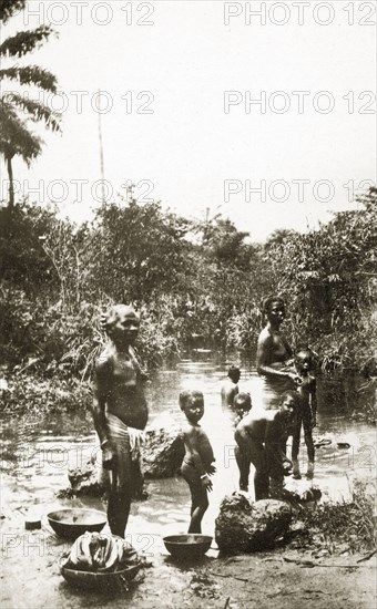 River laundry. A group of half-naked African women and children wash clothes in a shallow river surrounded by overgrown vegetation. Eastern Africa, 1-17 January 1924., Eastern Africa, Africa.