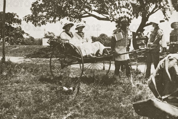 Rickshaw puller in Durban. A rickshaw puller wearing traditional costume and a flamboyantly feathered headdress gives a ride to two European men dressed in white naval uniforms. Durban, Natal (KwaZulu Natal), South Africa, 1-5 January 1924. Durban, KwaZulu Natal, South Africa, Southern Africa, Africa.