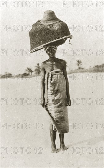 East African fisherwoman. An east African fisherwoman balances a large basket and fishing net on her head, topped off with a gentlemen's hat. Eastern Africa, 12-17 January 1924., Eastern Africa, Africa.