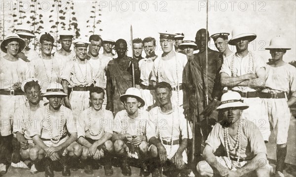 Sailors from HMS Delhi. A group of European sailors from HMS Delhi line up for a photograph with two east African hunters. Although uniformed, the sailors appear casual, wearing long strings of shell beads around their necks and smoking pipes. Eastern Africa, 12-17 January 1924., Eastern Africa, Africa.