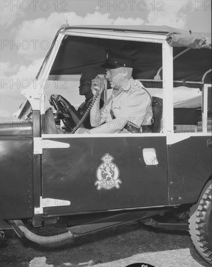 Kenyan police jeep. A uniformed policeman sits inside a police jeep operating a radio receiver during a royal visit. The insignia of the Kenya Police is displayed on the door of the vehicle, with the organisations' motto, 'Salus Populi', displayed beneath. Kenya, 10 September 1956. Kenya, Eastern Africa, Africa.