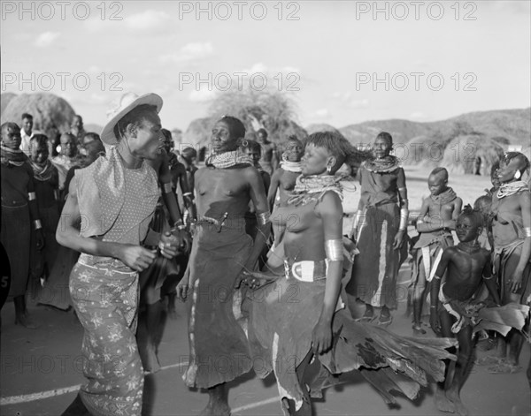 A Turkana 'ngoma'. A crowd of onlookers watch a Turkana man and two Turkana women wearing traditional dress and jewellery perform a 'ngoma'. Wamba, Kenya, 13 October 1956. Wamba, Rift Valley, Kenya, Eastern Africa, Africa.