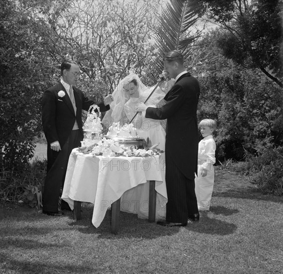 Cutting the cake with a ceremonial sword. The newlywed Williams and McLaughlin couple cut their wedding cake with a ceremonial sword. Kenya, 22 August 1956. Kenya, Eastern Africa, Africa.
