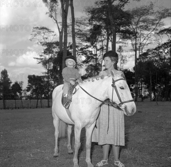 Children's pony riding. A woman smiles at a small child who sits proudly astride a white pony holding the reins in the class four competition at the SJAK show (Sports Journalists Association of Kenya). Kenya, 25 August 1956. Africa.