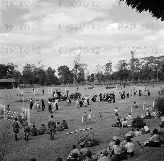 Square dancing at the SJAK show. A group of square dancers perform to a scattered audience in the arena at the SJAK show (Sports Journalists Association of Kenya) that contains a number of horse jumps. Kenya, 25 August 1956. Kenya, Eastern Africa, Africa.