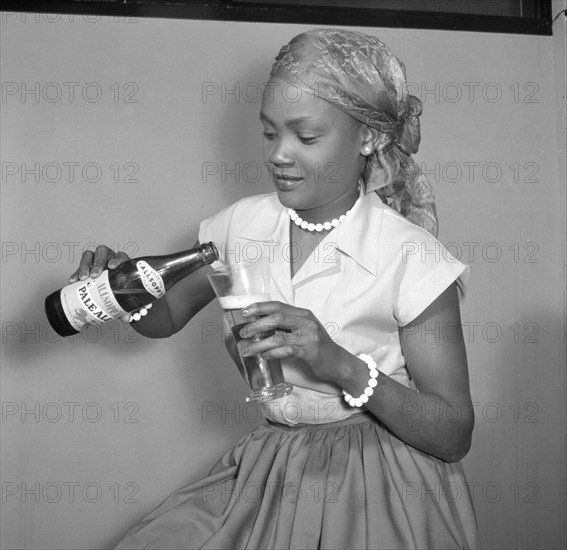 Promotional shot for Allsopp's Pale Ale. Promotional shot for Allsopp's Pale Ale. A smiling African girl wearing a headscarf, beads and Western-style clothing, pours a bottle of Allsopp's Pale Ale into a tall glass, label towards the camera. Kenya, 25 September 1956. Kenya, Eastern Africa, Africa.