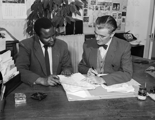 Authors work on a book. Promotional shot for the East African Literature Bureau. Two authors wearing suits, one African and one European, work together on a new book. Kenya, 25 September 1956. Kenya, Eastern Africa, Africa.