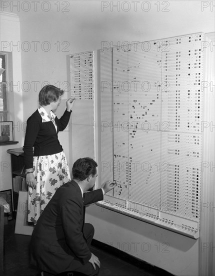 Wall chart at the Literature Bureau. Promotional shot for the East African Literature Bureau. Employees plot coloured markers onto sections of a large wall chart. Kenya, 25 September 1956. Kenya, Eastern Africa, Africa.