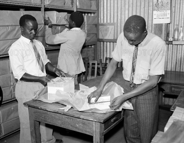 Packing books for dispatch. Promotional shot for the East African Literature Bureau. Three African men wearing shirts and ties wrap bundles of new books in paper and shelve them ready for dispatch. Kenya, 25 September 1956. Kenya, Eastern Africa, Africa.