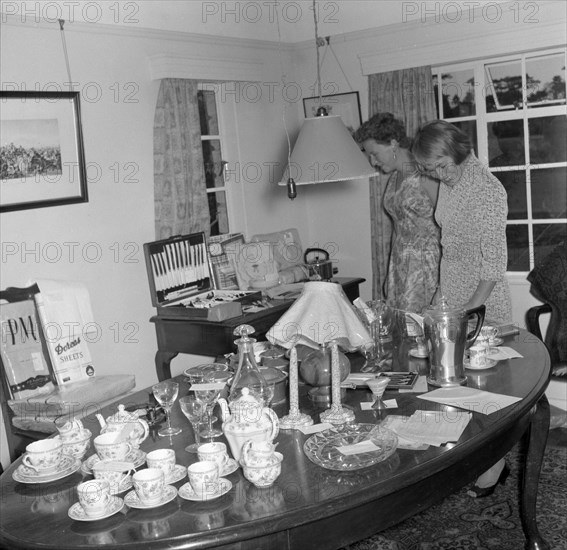 Inspecting the wedding presents. Two women examine wedding presents on display at the Patterson wedding. Amongst other items, the gifts include candlesticks, a tea set, a coffee jug, wine glasses, a decanter, a lamp shade, plates and a cutlery set. Kenya, 29 September 1956. Kenya, Eastern Africa, Africa.