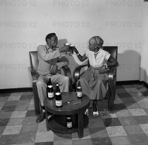 A toast to Allsopp's Pale Ale. Promotional shot for Allsop's Pale Ale. An African man and woman wearing Western clothes toast a drink to each other. Four bottles of Allsopp's Pale Ale and a bottle opener sit on the table in front of them. Kenya, 2 October 1956. Kenya, Eastern Africa, Africa.