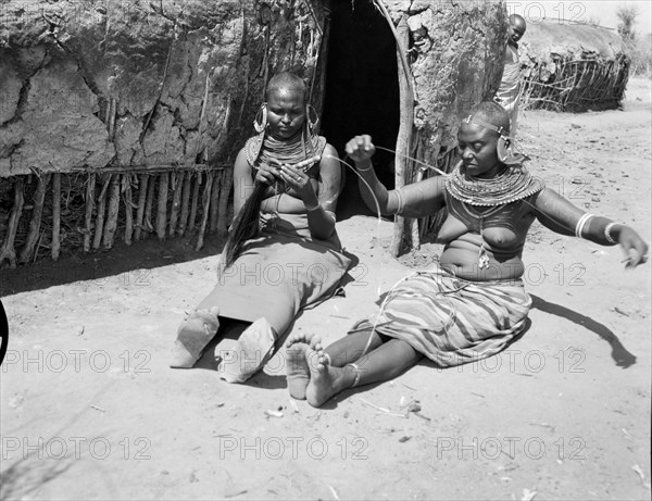 Samburu women threading beads. Two Samburu women sit outside a mud hut threading beads onto long wires. Naked from the waist up, they are decorated with heavily beaded necklaces and striking, ornate earrings. Wamba, Kenya, 13 October 1956. Wamba, Rift Valley, Kenya, Eastern Africa, Africa.