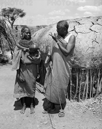 Lesanguniguni and his wife. A Samburu man identified as 'Lesanguriguri' chats to his young wife who holds a baby. Both wear traditional dress: her's decorated with striking, ornate jewellery. Wamba, Kenya, 13 October 1956. Wamba, Rift Valley, Kenya, Eastern Africa, Africa.