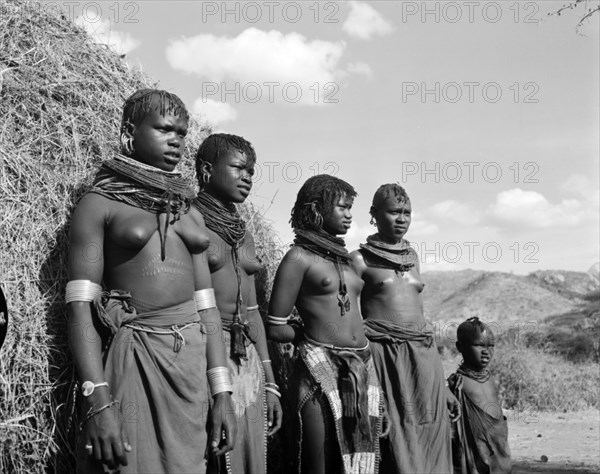 Turkana village girls. Portrait of four young Turkana women and a child outside a thatched village hut. Semi-naked, they display scarification marks on their bellies and wear traditional jewellery including heavily beaded necklaces, armlets and numerous hooped earrings. Wamba, Kenya, 13 October 1956. Wamba, Rift Valley, Kenya, Eastern Africa, Africa.