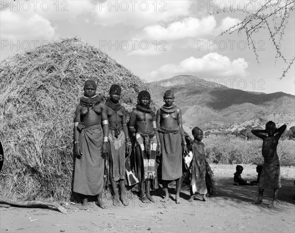 Turkana village girls. Four young Turkana women and children pictured outside a thatched village hut. They are semi-naked and wear traditional jewellery including heavily beaded necklaces, armlets and numerous hooped earrings. Wamba, Kenya, 13 October 1956. Wamba, Rift Valley, Kenya, Eastern Africa, Africa.