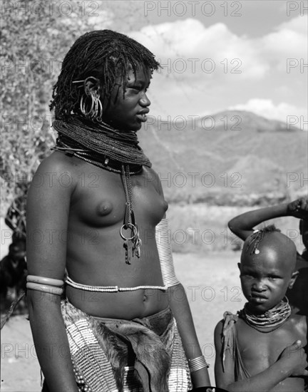 A young Turkana woman. Portrait of a young Turkana woman with a child in shot. Semi-naked, she displays scarification marks on her belly and is decorated with traditional jewellery including a heavily beaded necklace and belt, armlets and numerous hooped earrings. Her hair is tightly braided and she wears a bunch of keys around her neck. Wamba, Kenya, 13 October 1956. Wamba, Rift Valley, Kenya, Eastern Africa, Africa.