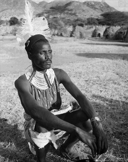 Portrait of a Turkana warrior. A Turkana warrior crouches on the ground. He wears traditional dress including a striking feathered hat and ornate beaded necklace. Wamba, Kenya, 13 October 1956. Wamba, Rift Valley, Kenya, Eastern Africa, Africa.