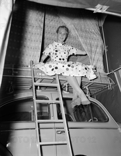 Girl in a spotted dress. A young woman in a spotted dress sits, ankles crossed, on a padded rack above a car at the Cooper Motors stand at the Royal Show. Kenya, 17 October 1956. Kenya, Eastern Africa, Africa.