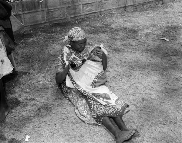 Pepsi cola baby. An African woman sits on the ground, feeding her baby wrapped in a cloth sling from a Pepsi Cola bottle. Kenya, 17-20 October 1956. Kenya, Eastern Africa, Africa.