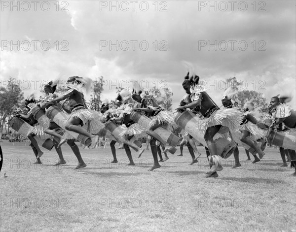 Chuka drummers at the Royal Show. A group of Chuka drummers perform a traditional dance at the Royal Show, their tubular drums tucked between their legs. They wear ceremonial dress including feathered headdresses and grass skirts. Kenya, 17-20 October 1956. Kenya, Eastern Africa, Africa.