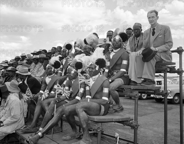 Turkana welcoming committee. Turkana men in ceremonial dress sit on a spectator stand amongst an African assembly awaiting the arrival of Princess Margaret. They wear elaborate headdresses and armbands and each holds a small union jack flag. Kenya, 18 October 1956. Kenya, Eastern Africa, Africa.