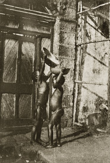 Young water carriers. A young child stretches up to pour water from one bowl into another balanced upon a friend's head. Ceylon (Sri Lanka), 27-31 January 1924. Sri Lanka, Southern Asia, Asia.