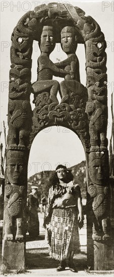 Beneath a Maori arch. A Maori woman in traditional dress stands beneath an ornate Maori arch that has been elaborately carved with wooden figures and swirling patterns. Rotorua, New Zealand, 10-17 May 1924. Rotorua, Bay of Plenty, New Zealand, New Zealand, Oceania.