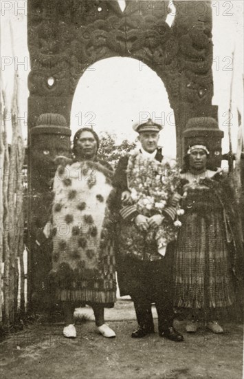 Rear Admiral Brand visits a Maori arch. Rear Admiral Sir Hubert Brand stands between two Maori women beneath an elaborately carved wooden arch. Whakarewarewa, New Zealand, 10-17 May 1924. Whakarewarewa, Bay of Plenty, New Zealand, New Zealand, Oceania.