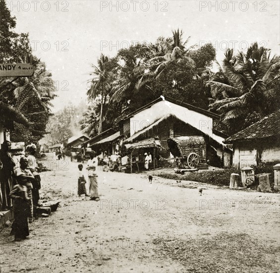 Ceylonian street scene. A wide dirt track flanked by low buildings and palm trees serves as the main road through a busy village. Signposts on the roadside indicate the distance to two other Ceylonian towns, Kandy, only four miles away and Colombo, 68 miles to the west. Peridenia, Ceylon (Sri Lanka), 27-31 January 1924. Peridenia, Central (Sri Lanka), Sri Lanka, Southern Asia, Asia.