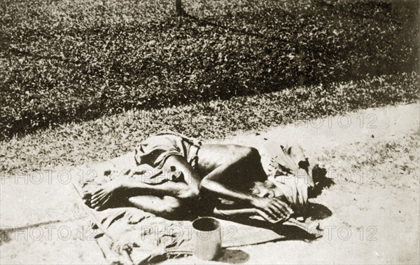 A India fakir in Ceylon. An Indian fakir lies hunched up with a begging bowl on a sack outside the sacred Dalada Maligawa or Temple of the Buddha's Tooth. Kandy, Ceylon (Sri Lanka), 27-31 January 1924. Kandy, Central (Sri Lanka), Sri Lanka, Southern Asia, Asia.