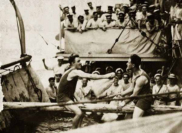Sports at sea. Two crew members aboard a British Special Service Squadron warship entertain their shipmates by trying to knock each other off a log suspended over water. Location unknown, circa 1924.