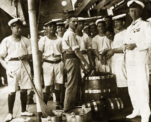 Grog and lime juice. Uniformed sailors aboard a British Special Service Squadron warship queue to be issued rations of 'grog and lime juice' from a barrel on deck. Location unknown, circa 1924.