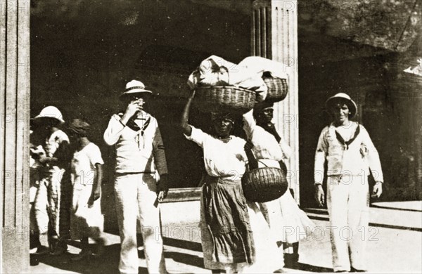 Jamaican street scene. Uniformed sailors from the British Special Service Squadron pose for the camera beside two Jamaican women carrying baskets of laundry on their heads. Kingston, Jamaica, 26-30 July 1924. Kingston, Kingston, Jamaica, Caribbean, North America .