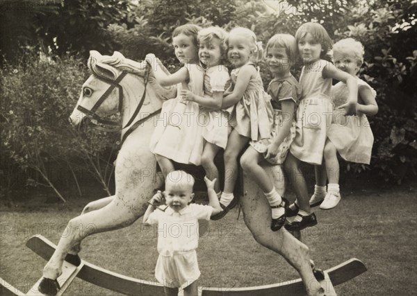 Alison's birthday party. Six smiling young girls sit astride a large rocking horse at 'Alison's birthday party' (Alison is pictured far right, the eldest daughter of Alan and Audrey Leonard). A young boy, unable to fit on the horse, holds onto its stirrup instead. Calcutta (Kolkata), India, circa 1954. Kolkata, West Bengal, India, Southern Asia, Asia.