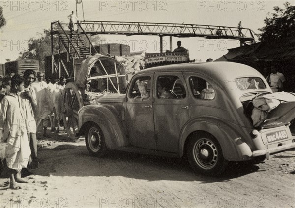 Journey to a dak bungalow. "David and Marjorie's car with Caroline at the window, held up in a wonderful confusion that had to be seen to be believed". A car queues amid the chaos of the busy railway stop, luggage spilling from its trunk, on the way to a dak bungalow (traveller's rest house). India, 1954. India, Southern Asia, Asia.