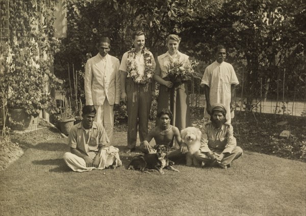 Members of the Leonard household. Alan and Audrey Leonard, adorned with flowers, pose for the camera with their team of Indian servants and two pet dogs in the garden of their colonial house. Calcutta (Kolkata), India, circa 1954. Kolkata, West Bengal, India, Southern Asia, Asia.