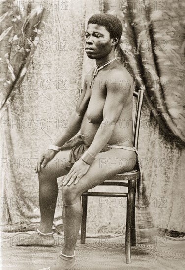 Seated woman with scarified stomach. Portrait of a seated woman, with scarified stomach and wearing an apron and back cloth around her waist and simple jewellery on her neck, wrists and ankles. Western Africa, circa 1917., Western Africa, Africa.