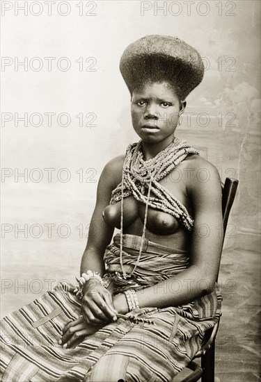 Seated woman with high hairstyle. Portrait of a young woman with an elaborately raised coiffure, seated, wearing a wrap skirt and beaded necklace-cum-bodice. Her dress is similar to that of contemporary studio photographs of Fante women. Western Africa, circa 1917., Western Africa, Africa.